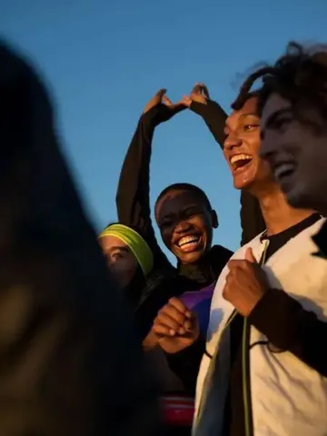 Four young people stand close together outdoors at sunset, one with hands raised overhead, backlit by warm light against a clear sky.