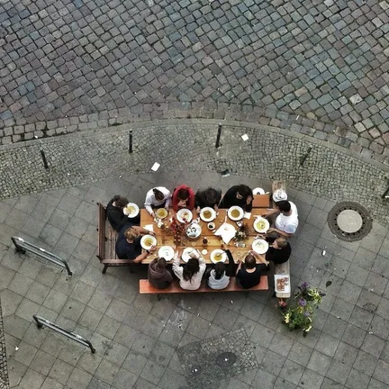 An overhead view shows a group of people seated at a long table set for a meal on a cobblestone square.