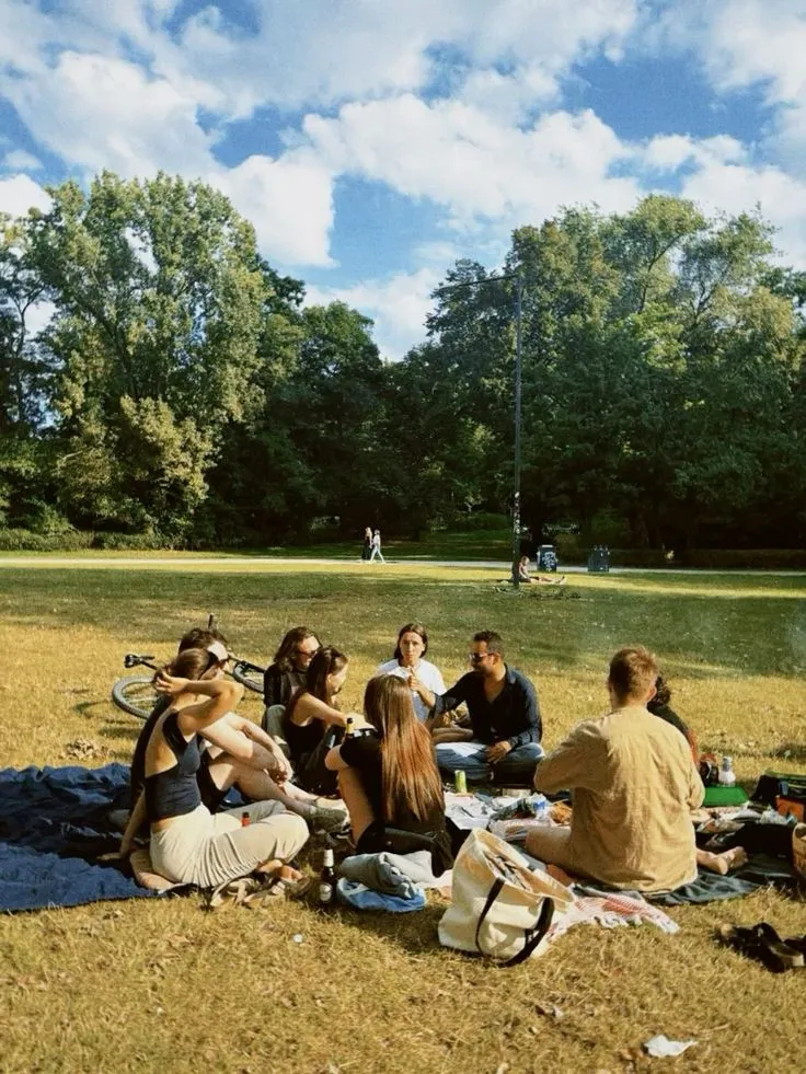 A group of people sit on blankets on grass in a park, talking together under a partly cloudy sky.