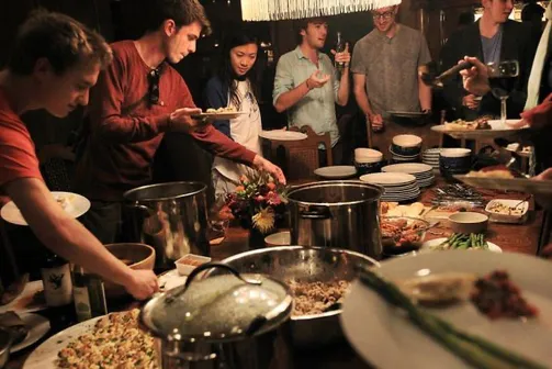 People serve themselves food from pots and platters set on a counter in a dimly lit kitchen.