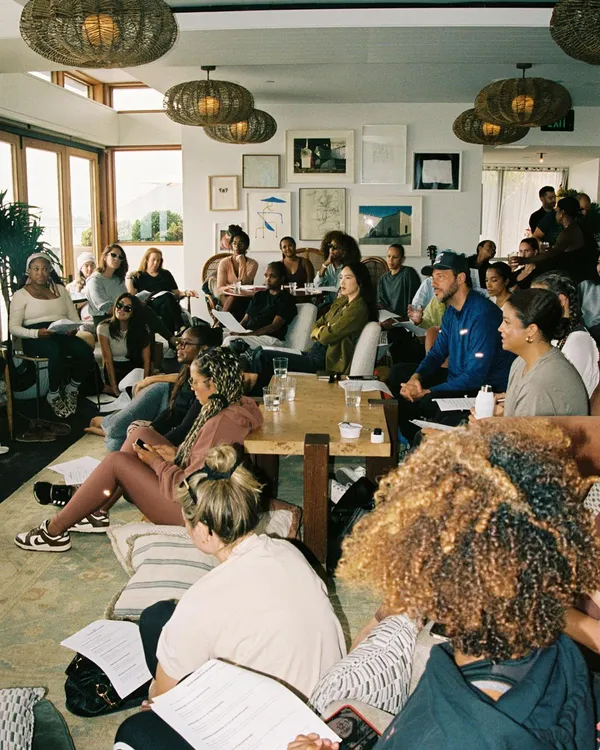 A large group sits and stands in a living room holding printed pages, with woven pendant lights hanging from the ceiling.