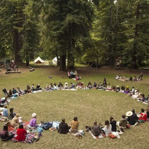 A large group sits in a circle on a grass clearing surrounded by tall trees, with tents visible in the background.