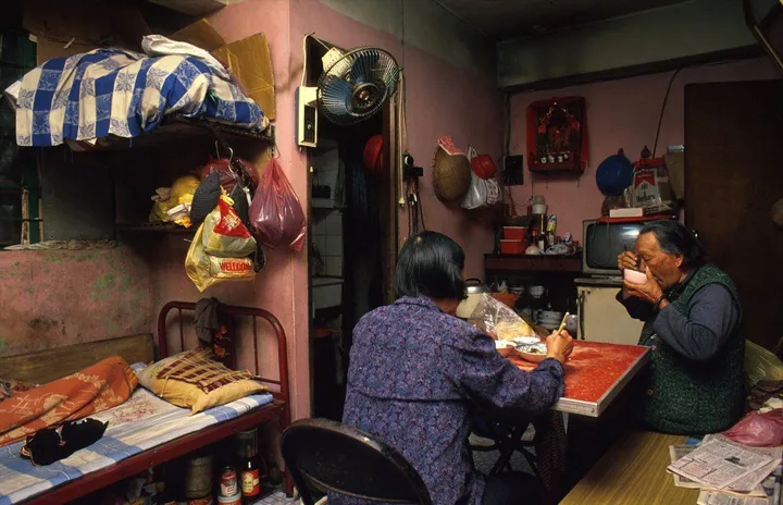 Two people sit at a small table in a cluttered room, one eating and one drinking from a cup.
